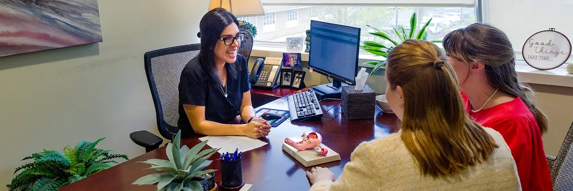 o Nurse practitioner Katherine Mendez seated at a desk, smiling while engaged in a consultation with a lesbian couple.